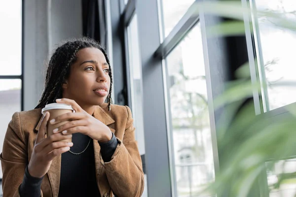 African american businesswoman holding coffee to go near window in office