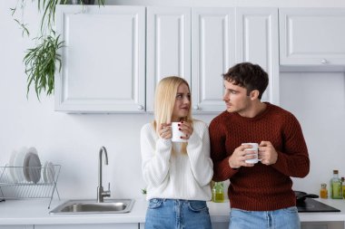 young couple in knitted sweaters looking at each other while holding cups with tea and warming up in winter clipart