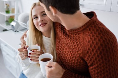 cheerful young couple in knitted sweaters hugging and holding cups with tea while warming up in winter clipart