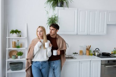 young couple in sweaters standing covered in blanket and holding cups of tea clipart