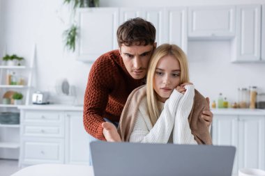 confused man in sweater and blonde woman looking at laptop while working from home clipart