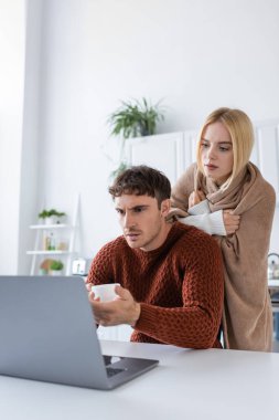 Sarışın kadın battaniyeye sarılıp erkek arkadaşının arkasında dizüstü bilgisayar kullanarak evde çalışıyor. 