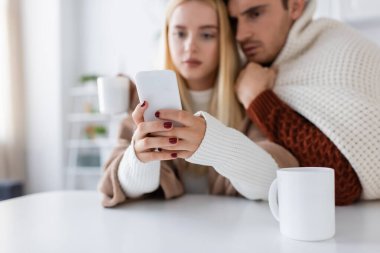 Erkek arkadaşının yanında akıllı telefon kullanan bulanık bir kadın ve masaya odaklanmış bir fincan çay.