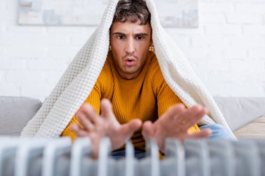 Duygusal genç adam battaniyeyle örtülmüş, ısıtıcının yanında ellerini ısıtıyor.
