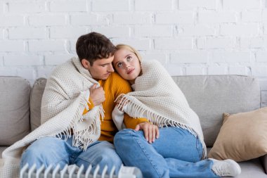 Genç çift battaniyeyle örtülmüş koltukta oturuyor ve modern radyatör ısıtıcısının yanında ısınıyor. 
