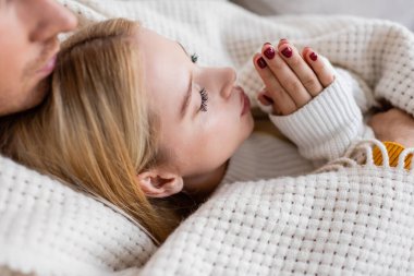 high angle view of blonde woman in white sweater warming up hands while lying with boyfriend covered in blanket  clipart