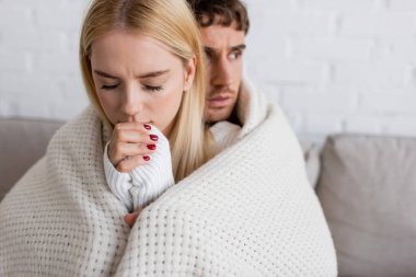 blonde woman in white sweater warming up hands near boyfriend covered in blanket  clipart