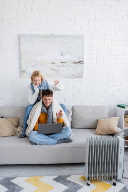 cheerful couple in scarfs and sweaters waving hands during video call on laptop  clipart