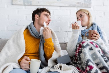 Kanepede otururken ellerinde çay ve mendillerle battaniyelere sarılı hasta çift.