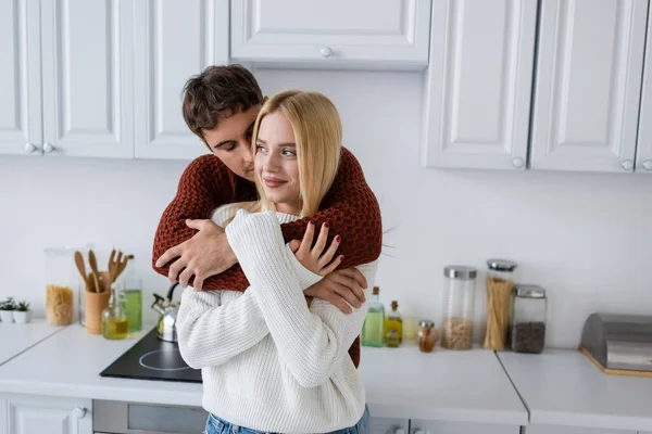 穿着红色毛衣的年轻人在厨房里高兴地拥抱着金发碧眼的女朋友 — 图库照片