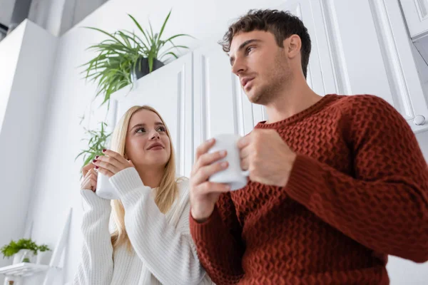 low angle view of young woman in knitted sweater holding cup of tea while looking at boyfriend