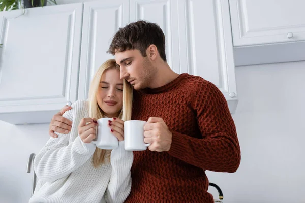 ニットのセーターを着た若いカップルがお茶を飲みながら抱き合ってカップを握って冬になると — ストック写真