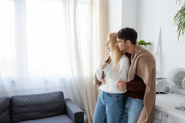 Jovem Casal Coberto Cobertor Abraçando Enquanto Segurando Xícaras Chá Casa — Fotografia de Stock