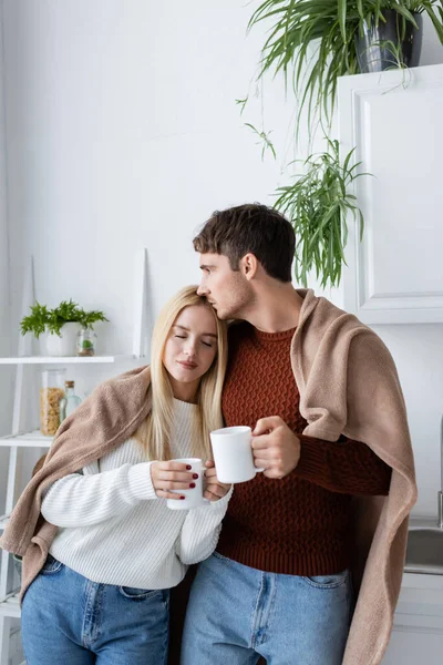 Jovem Casal Coberto Cobertor Abraçando Enquanto Segurando Xícaras Chá Casa — Fotografia de Stock