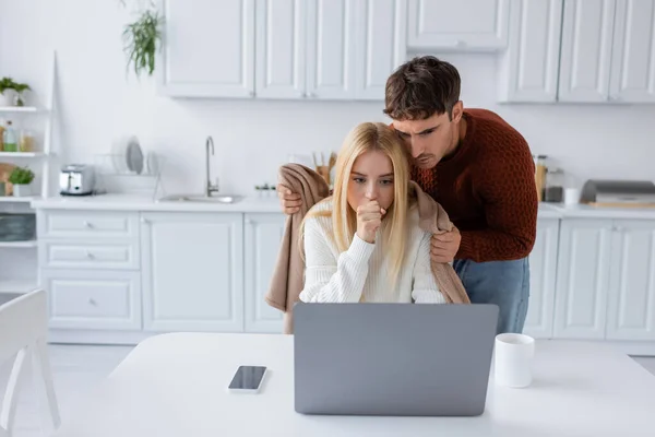 头戴毛衣的男人拿着毛毯 靠近紧张的女朋友 在家里用笔记本电脑干活 — 图库照片