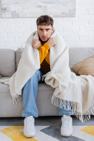 frustrated young man covered in blanket sitting on sofa while getting warm in living room 