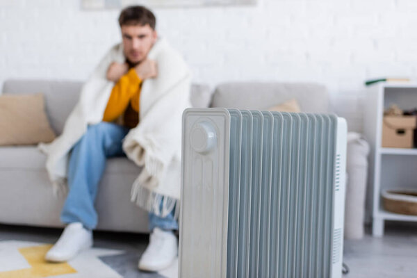 radiator heater near blurred man covered in blanket sitting on sofa in living room 