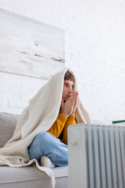 young man covered in blanket sitting on sofa and warming up hands near radiator heater