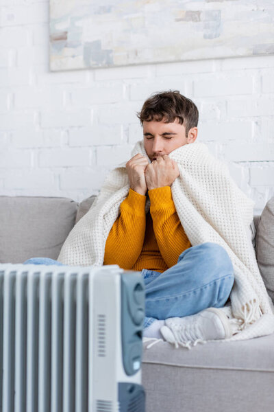 young man with closed eyes holding blanket and sitting on sofa near radiator heater in winter 