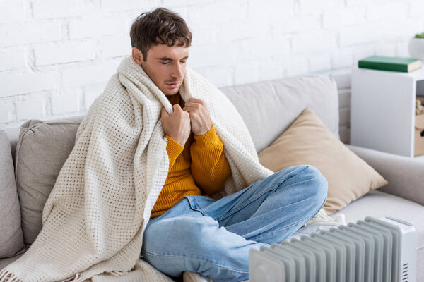 young man with closed eyes holding blanket while sitting on sofa near radiator heater in winter 
