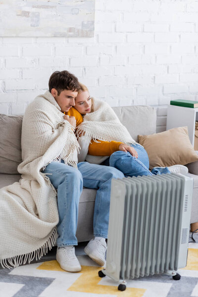young couple covered in blanket sitting on sofa and warming near modern radiator heater 