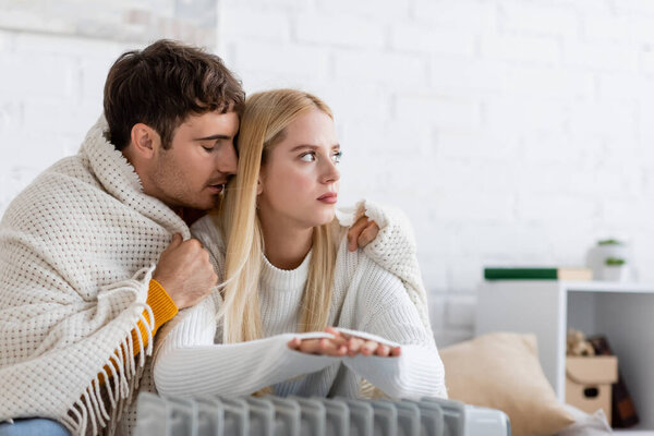 young couple covered in blanket warming near modern radiator heater 