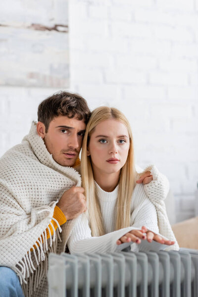 young couple covered in blanket warming near modern radiator heater at home