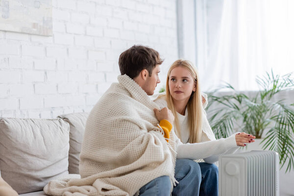 young couple covered in blanket sitting on couch and warming near radiator heater at home