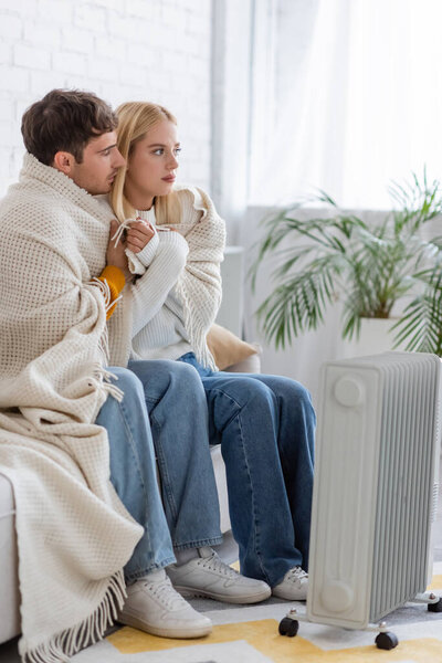 young couple covered in warm blanket sitting on couch near radiator heater at home