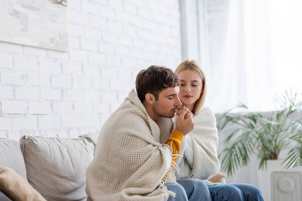 young man warming hand of girlfriend covered in blanket sitting in living room