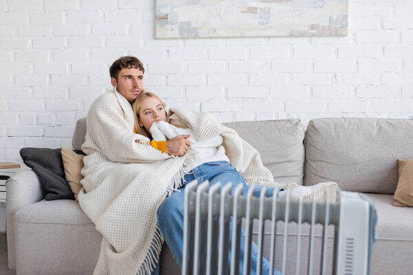 young couple covered in warm blanket hugging on couch near radiator heater at home 