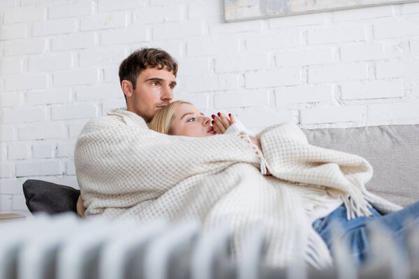 young couple covered in warm blanket hugging on couch near blurred radiator heater at home 