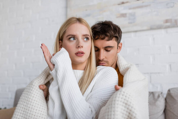 caring man covering blonde girlfriend in white sweater with blanket at home 