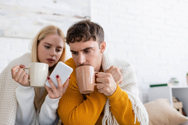 young woman holding cup of tea and smartphone near boyfriend covered with blanket 