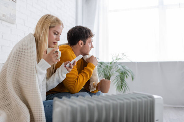 young woman with cup of tea using smartphone near boyfriend covered in blanket while sitting near radiator heater