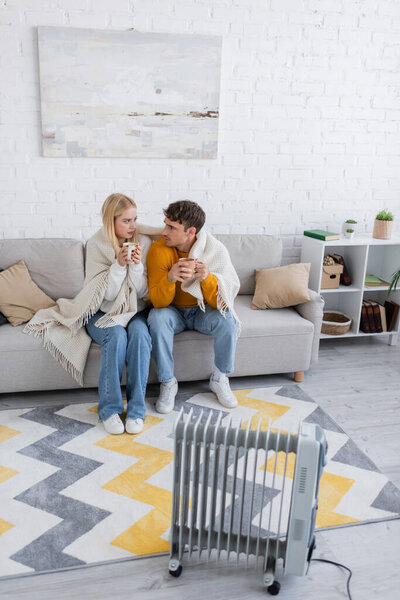 young couple covered in blanket sitting with cups of tea near radiator heater
