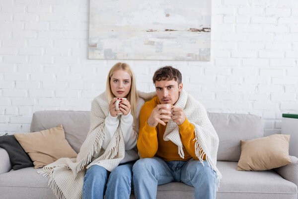 young couple covered in blanket sitting on couch and holding cups of tea 