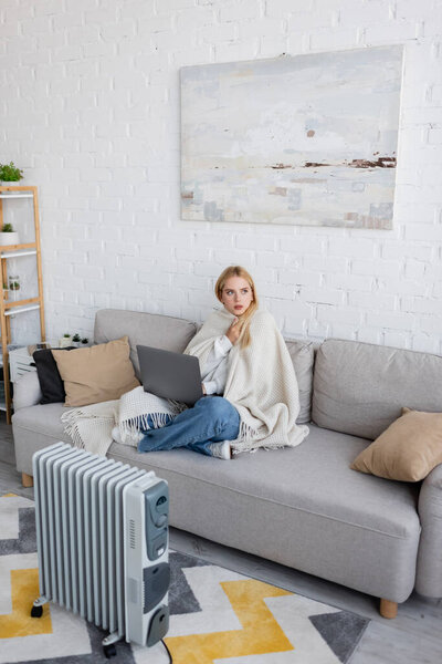 young blonde woman covered in white blanket using laptop while sitting on sofa near radiator heater