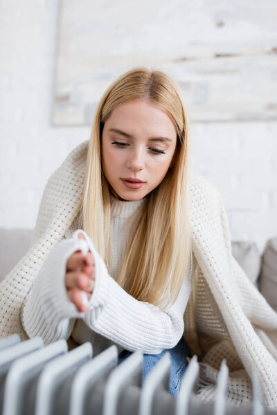 young blonde woman covered in blanket warming frozen hands near radiator heater