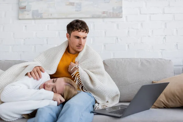 年轻女人穿着毛毯躺在男朋友的腿上睡觉 一边在家里工作一边看笔记本电脑 — 图库照片