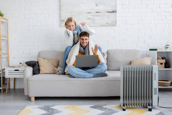 happy young couple in scarfs and sweaters waving hands during video call on laptop 
