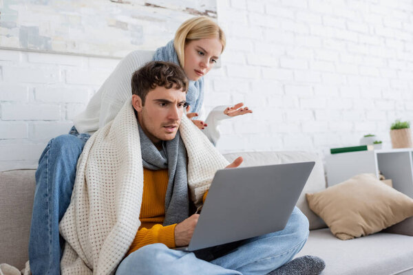 displeased blonde woman in sweater and scarf sitting near boyfriend and gesturing while looking at laptop 