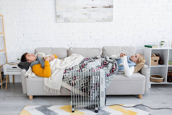 young man and blonde woman holding cups and lying under blankets on sofa near radiator heater