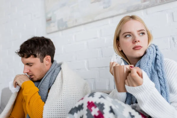 diseased blonde woman in scarf holding cup of tea and tissue near man coughing in living room