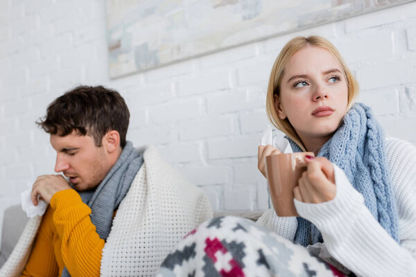 diseased blonde woman in scarf holding cup of tea and tissue near man coughing in living room 