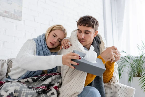 blonde woman in scarf holding tissue box near diseased boyfriend with cup of tea