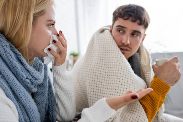 sick blonde woman in scarf sneezing in tissue near diseased boyfriend with cup of tea