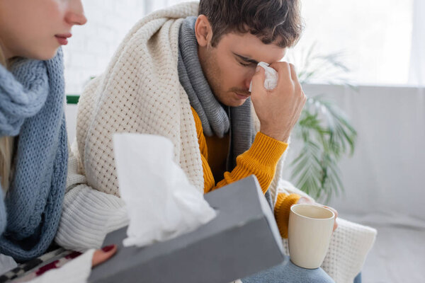 sick man with closed eyes holding napkin and cup near girlfriend with tissue box 
