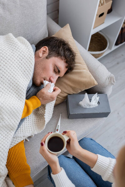 top view of woman holding cup of tea near sick boyfriend lying near tissue box 