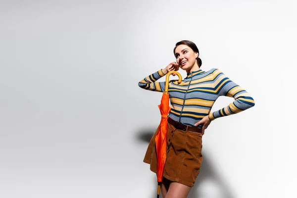 smiling young woman in striped turtleneck and skirt posing with hand on hip and holding umbrella on grey